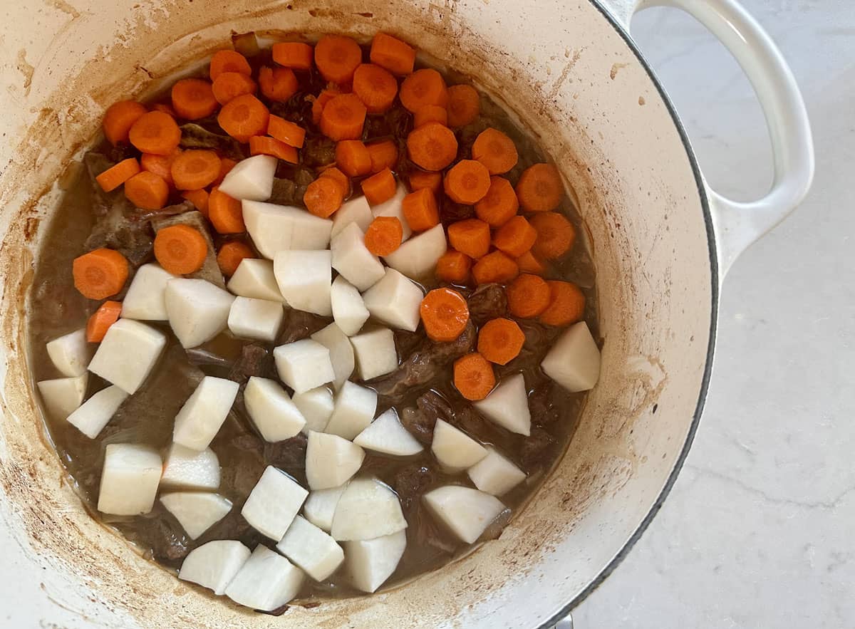 A white pot filled with chunks of carrots and turnips on top of browned meat in a stew, all set on a light-colored countertop.