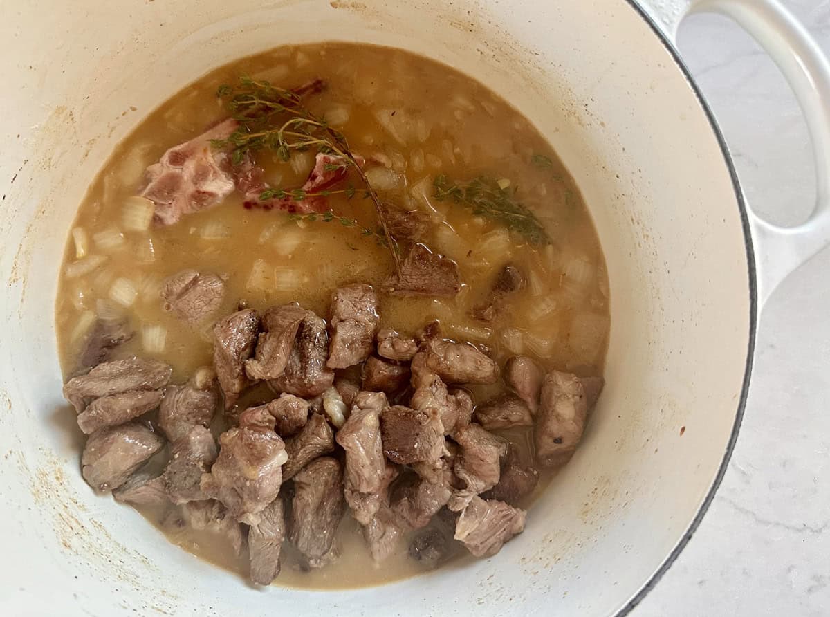 Chunks of browned meat, onions, herbs, and a bone simmer in a light broth inside a white Dutch oven on a light countertop.