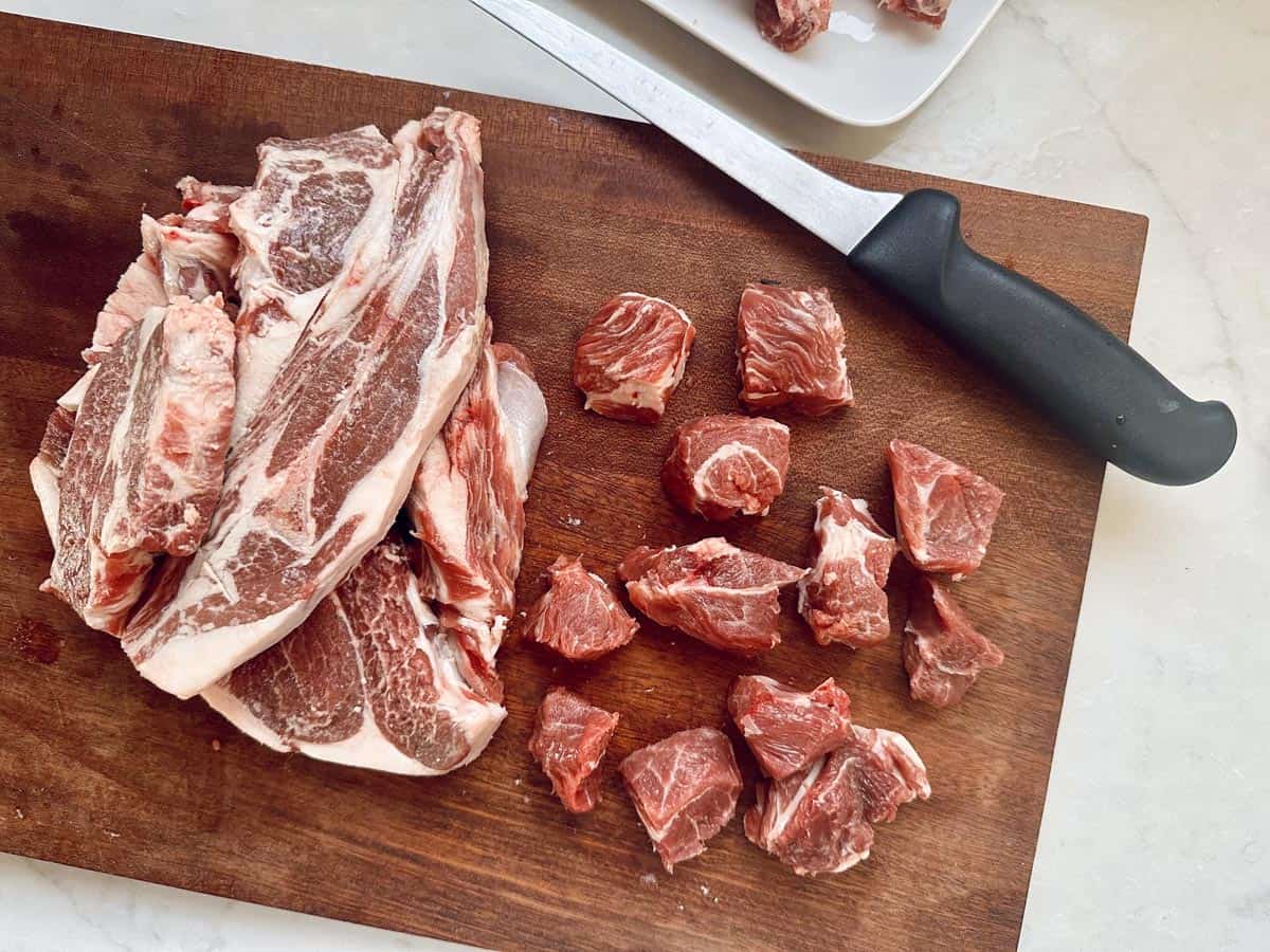 Raw beef strips and diced beef pieces on a wooden cutting board with a kitchen knife. A plate with more beef is partially visible in the background on a light countertop.
