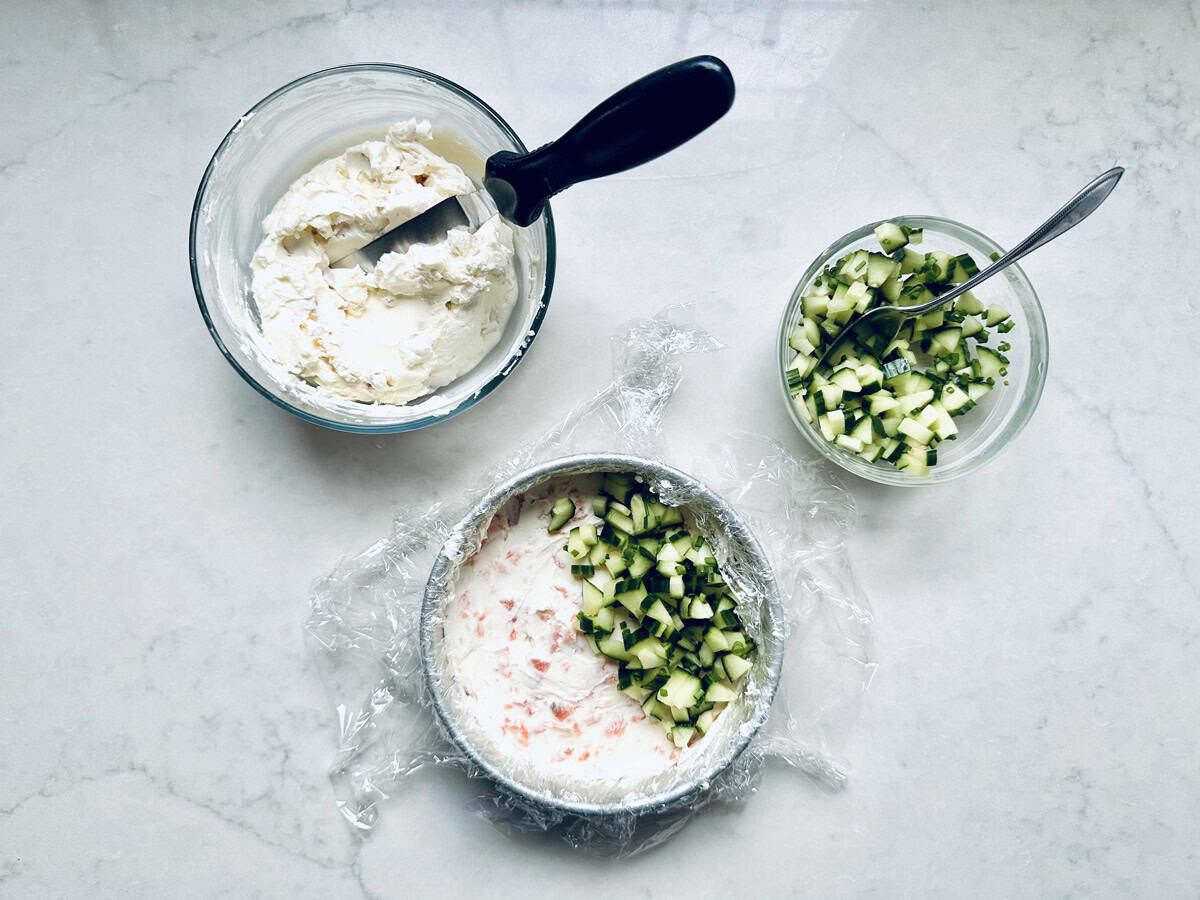Three bowls on a white surface: one with mixed cream cheese and a spatula, one with chopped cucumber and a spoon, and one with both ingredients layered in plastic wrap, ready to assemble into a delicious Gravlax Torta.