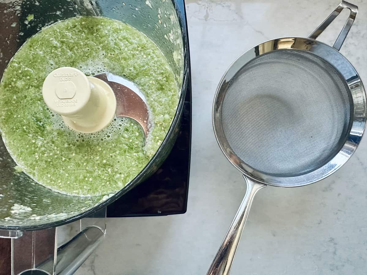 A food processor with a green, blended mixture sits next to a metal fine-mesh strainer on a light-colored countertop.