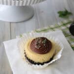 A mini cheesecake in a white paper liner with a chocolate cookie crust, topped with a swirl of chocolate frosting and a sprinkle of green garnish, sits on a napkin. More cheesecakes are displayed on a stand in the background.