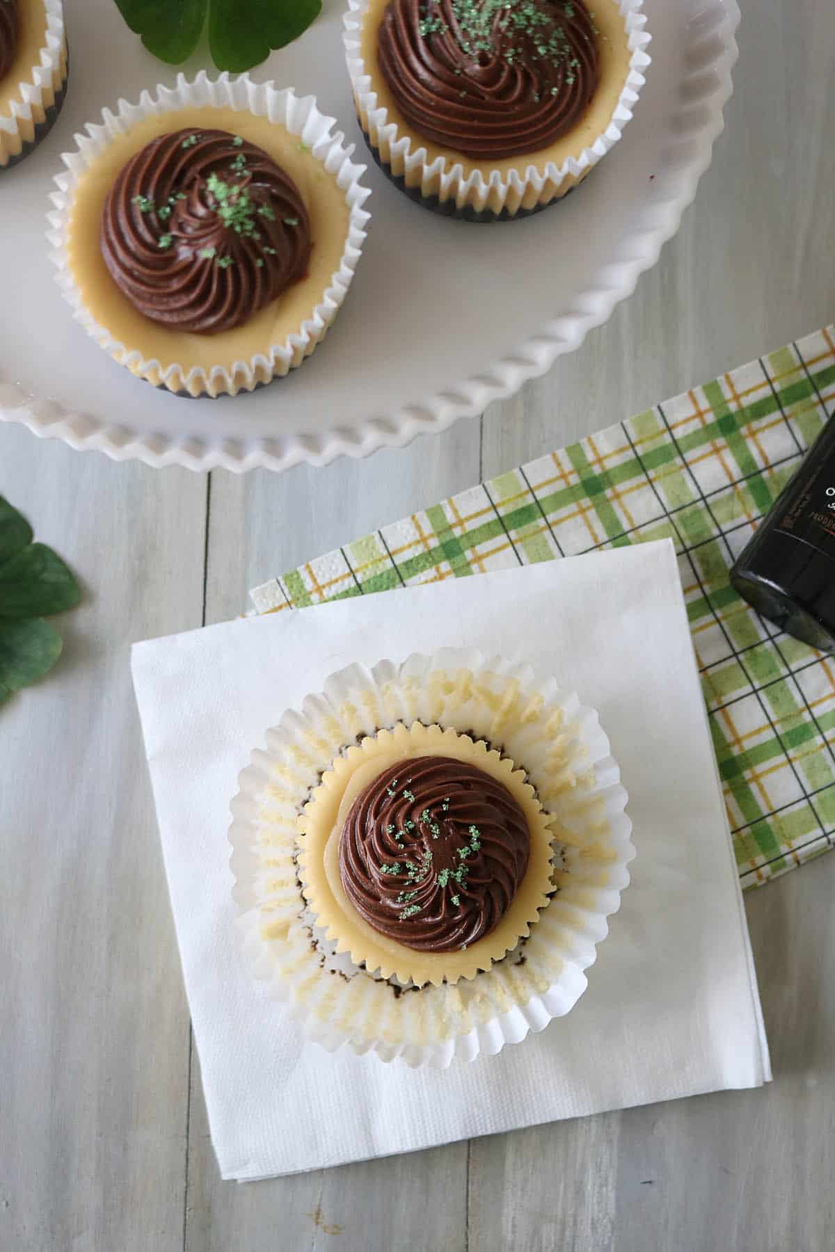 A cupcake with chocolate frosting and green sprinkles sits on a white napkin, with more cupcakes on a white stand nearby, atop a gray wooden table with a green and yellow plaid cloth.