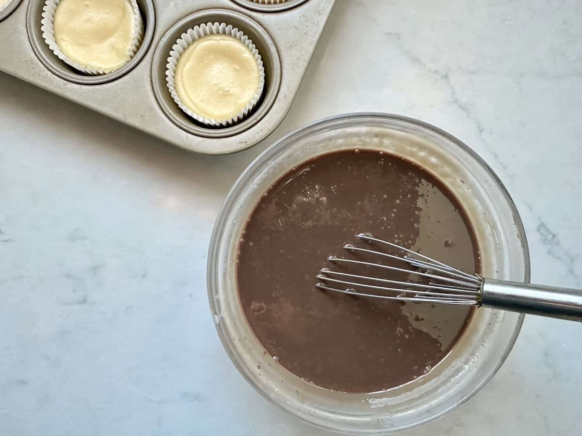 A glass bowl filled with chocolate batter and a metal whisk sits on a marble countertop next to a muffin tin with three unbaked cupcakes in paper liners.