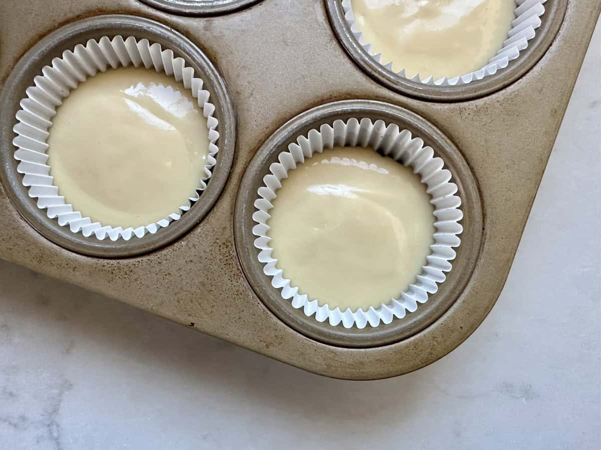 A muffin tin with white paper liners filled with pale, smooth batter, ready to be baked, sits on a light-colored surface.