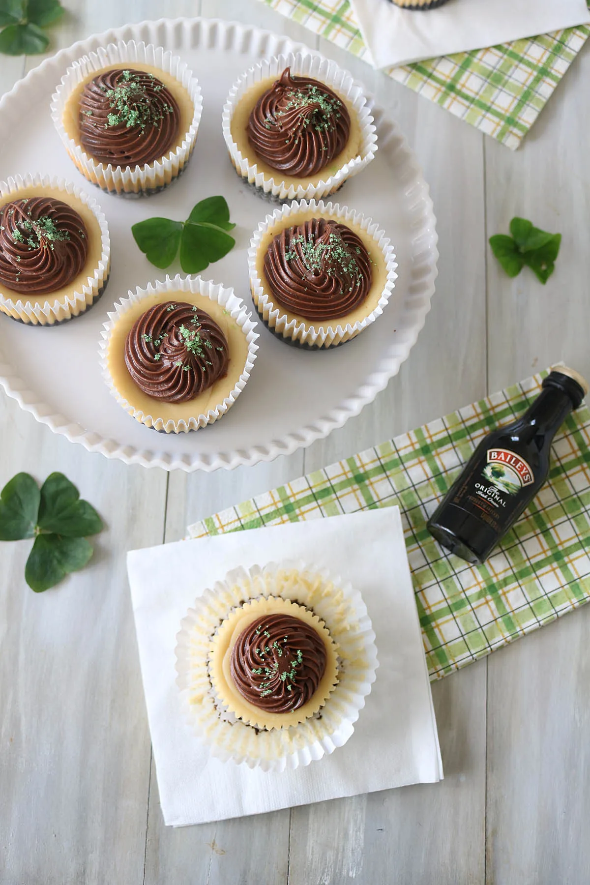 Cupcakes with chocolate swirled frosting and green sprinkles are displayed on a white plate and napkin. A small Baileys Irish Cream bottle and green shamrock leaves are set beside them on a light wooden surface.