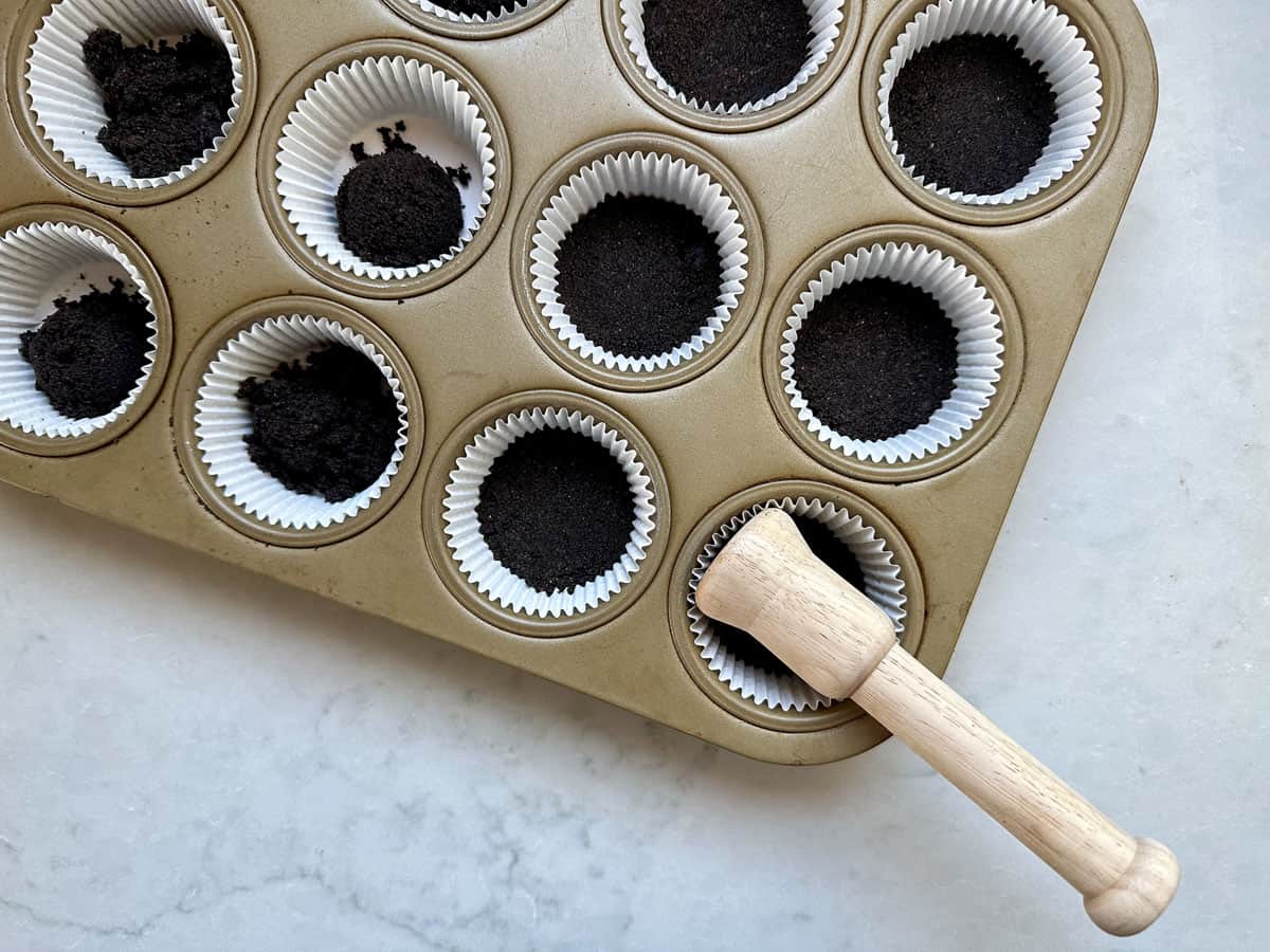 A muffin tin lined with white paper cups, some filled with a dark crumb mixture. A wooden tamper tool is in one cup, pressing the crumbs down. The tin is on a light countertop.