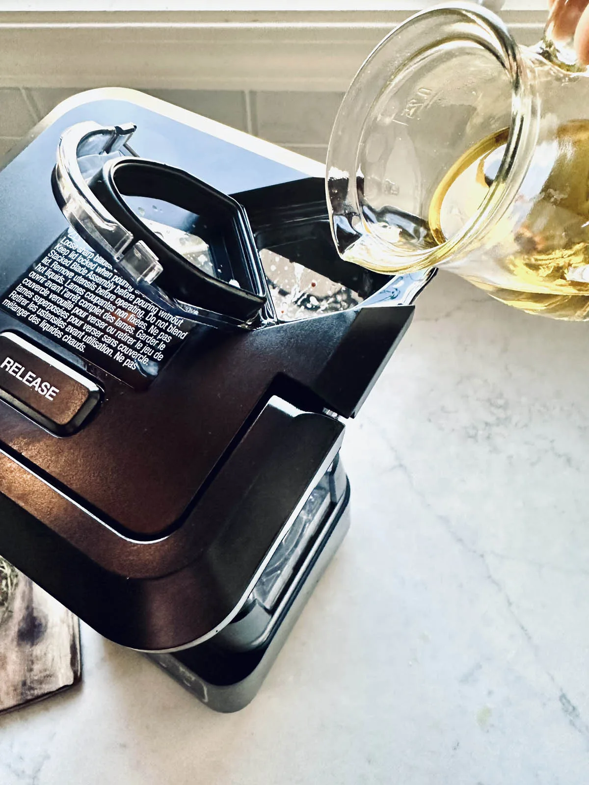 A hand pours oil from a glass container into a blender on a kitchen counter, with the blender lid partially open and recipe instructions visible on the lid.