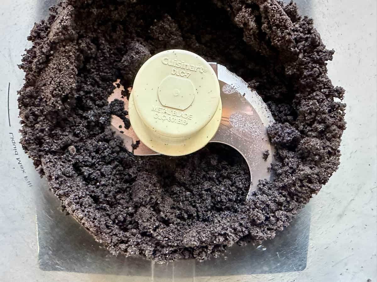 A food processor filled with finely ground chocolate cookie crumbs, forming a dark, crumbly mixture around the metal blade in the center.