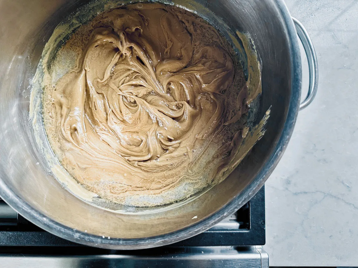 A large stainless steel pot sits on a stove, filled with a light brown, creamy mixture being stirred, possibly the beginning stages of making a roux or sauce. The countertop and stove edge are visible.