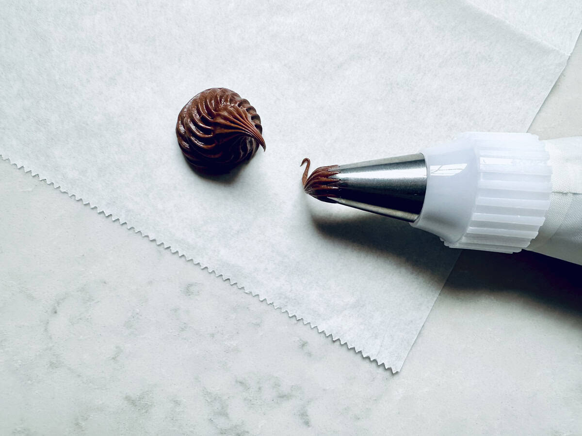 A piping bag with a metal tip is squeezing a swirl of chocolate frosting onto a piece of parchment paper on a light-colored surface.