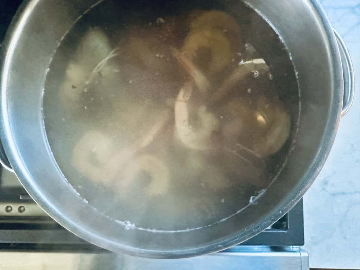 A large metal pot filled with water sits on a stove, with vegetables and possibly bones visible through the steam as they simmer, suggesting soup or broth is being made.