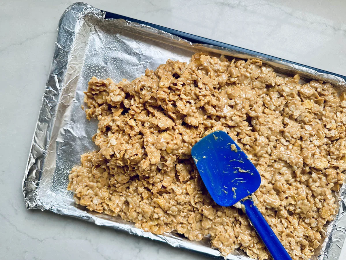 A baking tray lined with foil holds a mixture of Rice Krispies treats being spread with a blue silicone spatula on a light countertop.