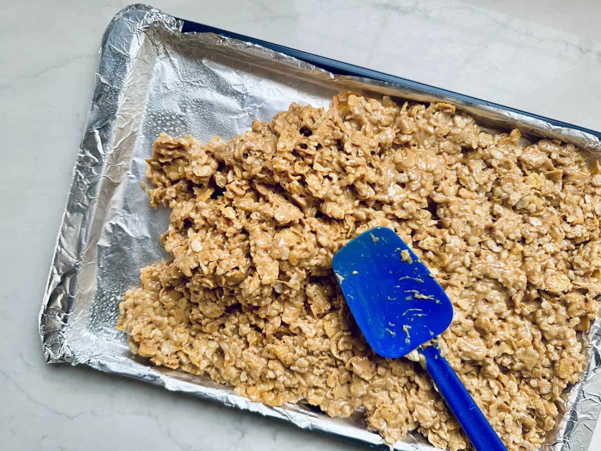 A baking tray lined with foil holds a mixture of Rice Krispies treats being spread with a blue silicone spatula on a light countertop.