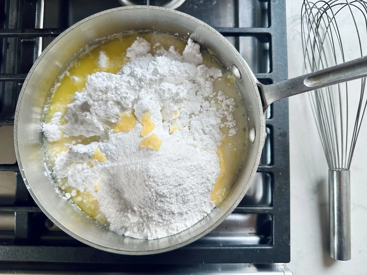 A saucepan on a stove contains melted butter and a large mound of powdered sugar. A metal whisk is placed on the countertop nearby.