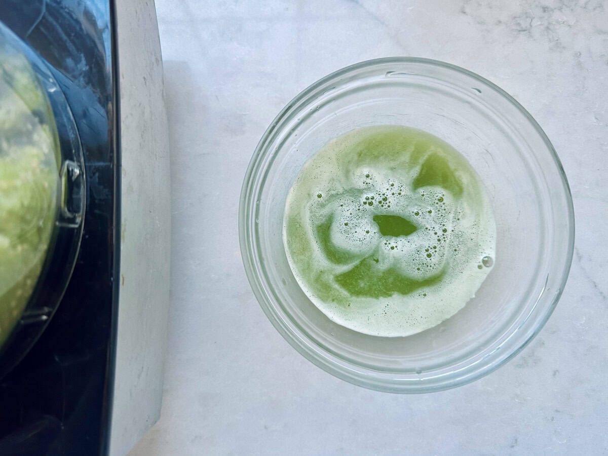 A glass filled with green juice sits on a white marble surface next to part of a blender with green remnants inside.