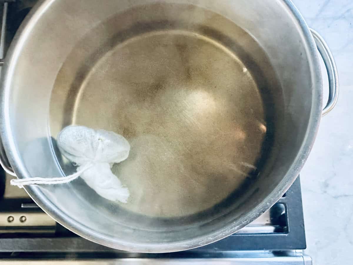 A large metal pot filled with hot water sits on a stovetop. Inside the pot, a small mesh bag tied with string floats near the surface. Steam is rising gently from the water.