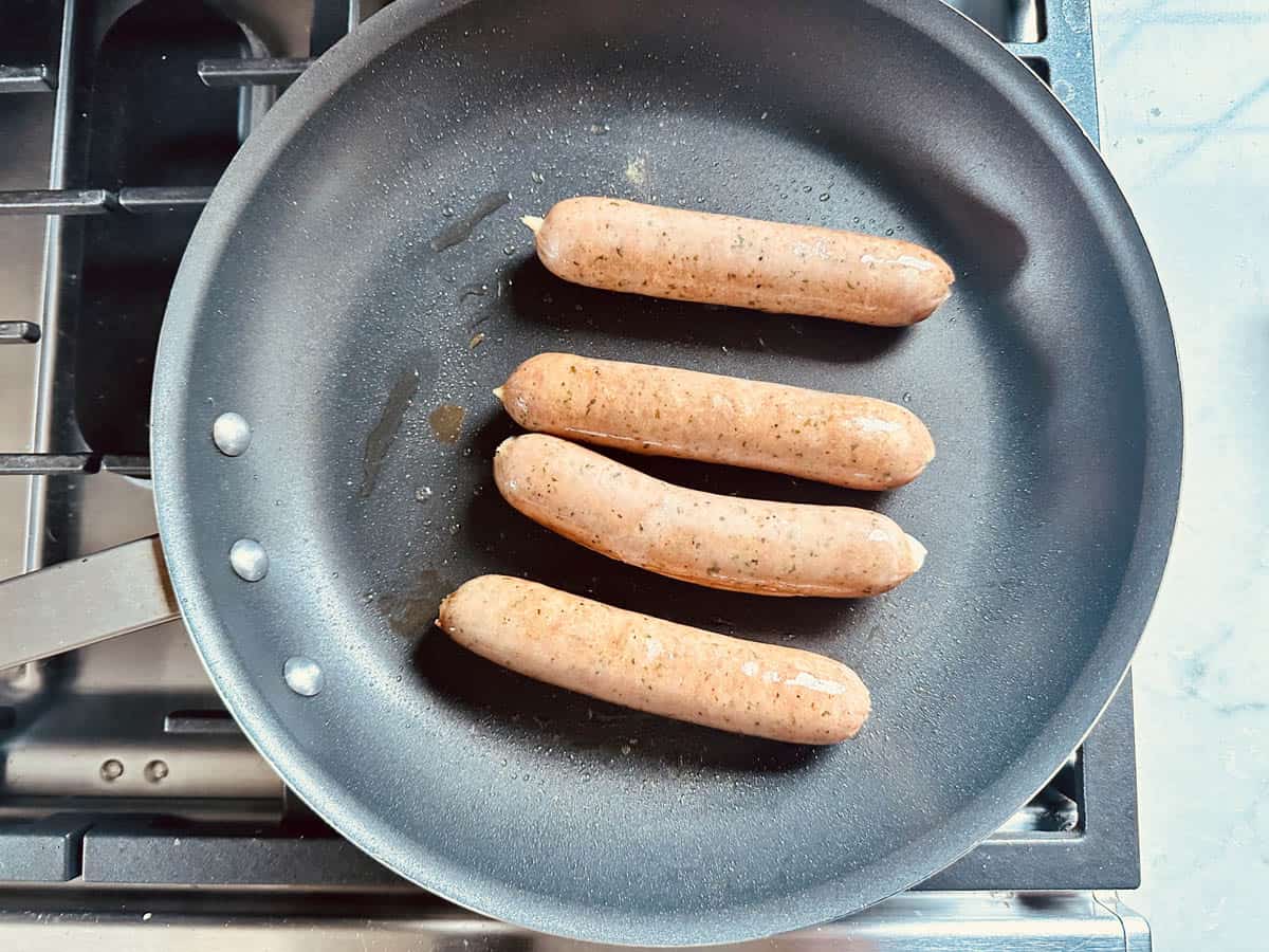 Four uncooked sausages are arranged in a nonstick frying pan on a stovetop, ready to be cooked for delicious Arugula Salad with Sausage and White Beans.