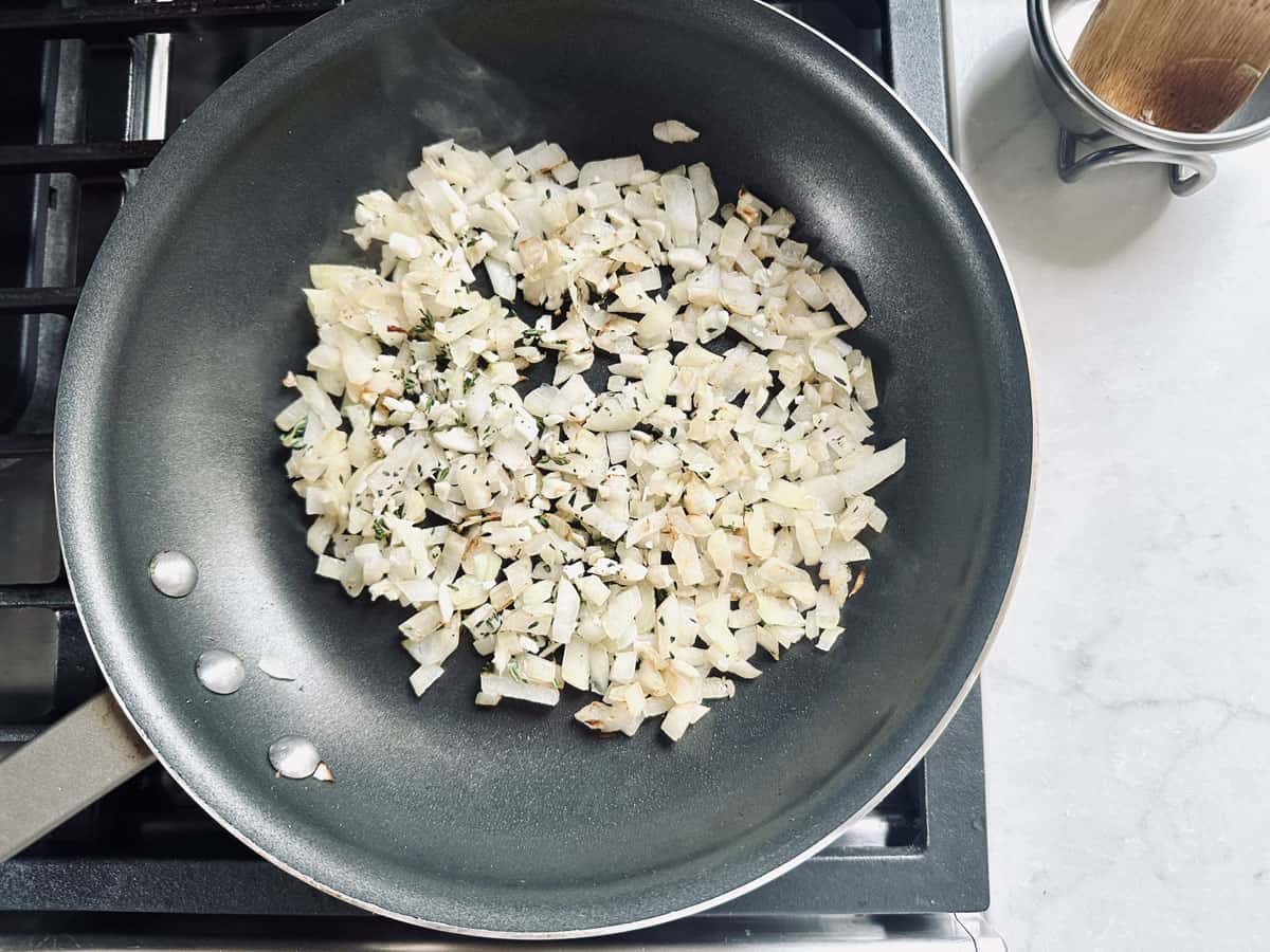 Chopped onions and herbs are being sautéed in a nonstick frying pan on a stovetop. A container with a wooden spoon lid is visible on the countertop nearby.