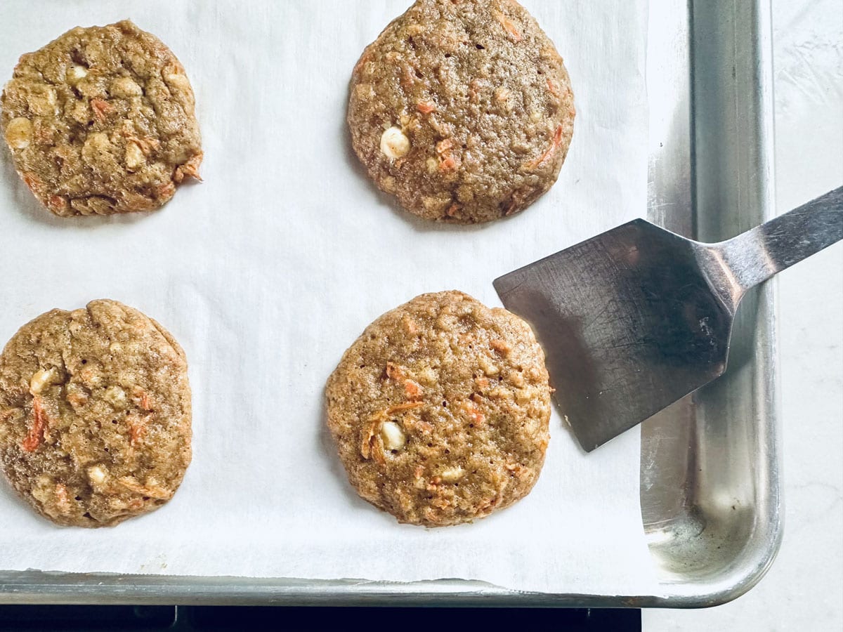 Four freshly baked cookies with visible white chocolate chips and shredded carrot on a parchment-lined baking sheet; a metal spatula is lifting one cookie.