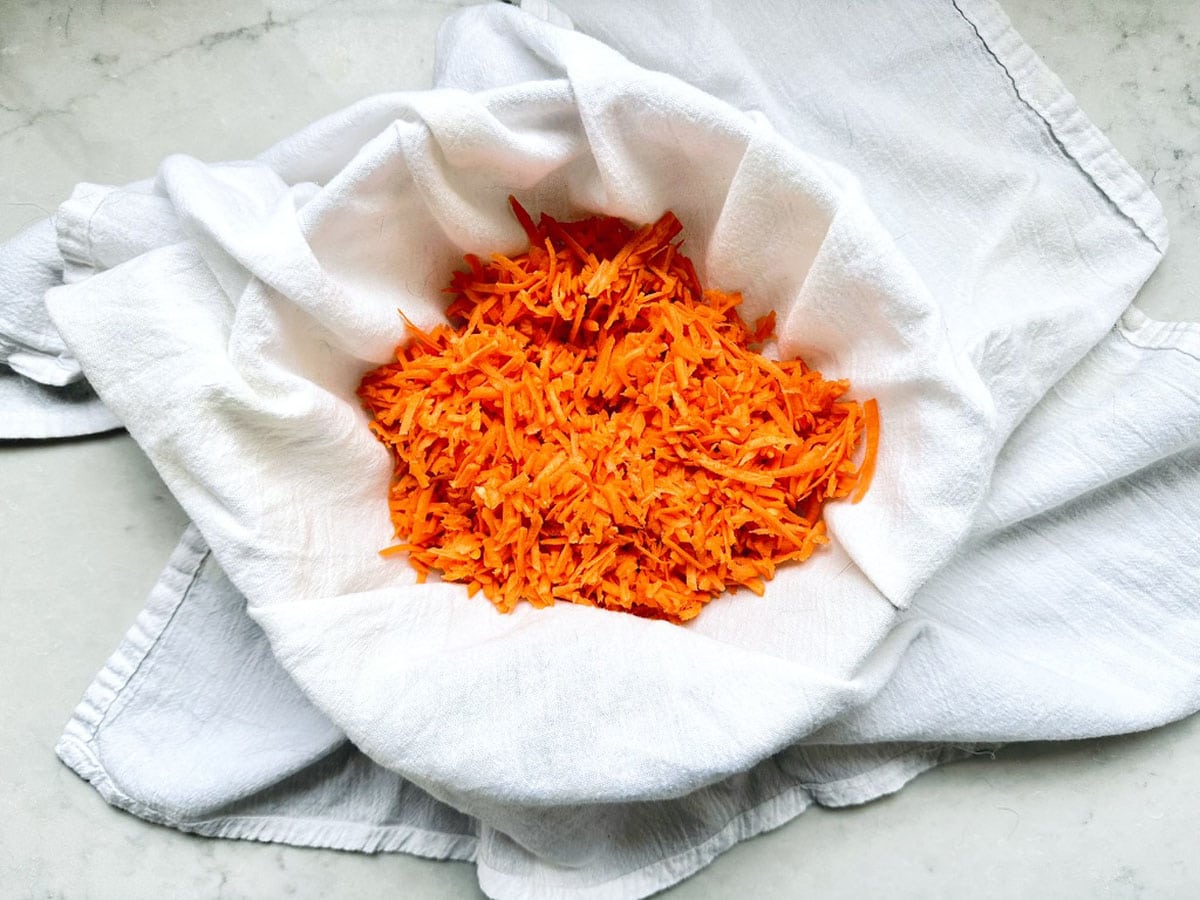 A white cloth-lined bowl filled with bright orange shredded carrots, resting on a light-colored surface.
