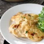 A white plate with two pieces of creamy baked fish, garnished with herbs, served alongside a small portion of fresh arugula greens. A skillet with more fish is visible in the background.