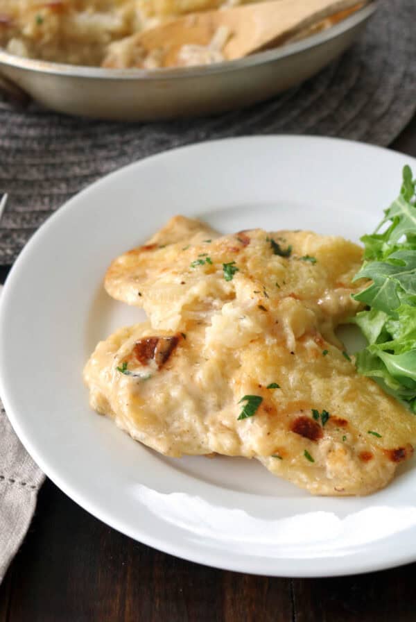 A white plate with two pieces of creamy baked fish, garnished with herbs, served alongside a small portion of fresh arugula greens. A skillet with more fish is visible in the background.
