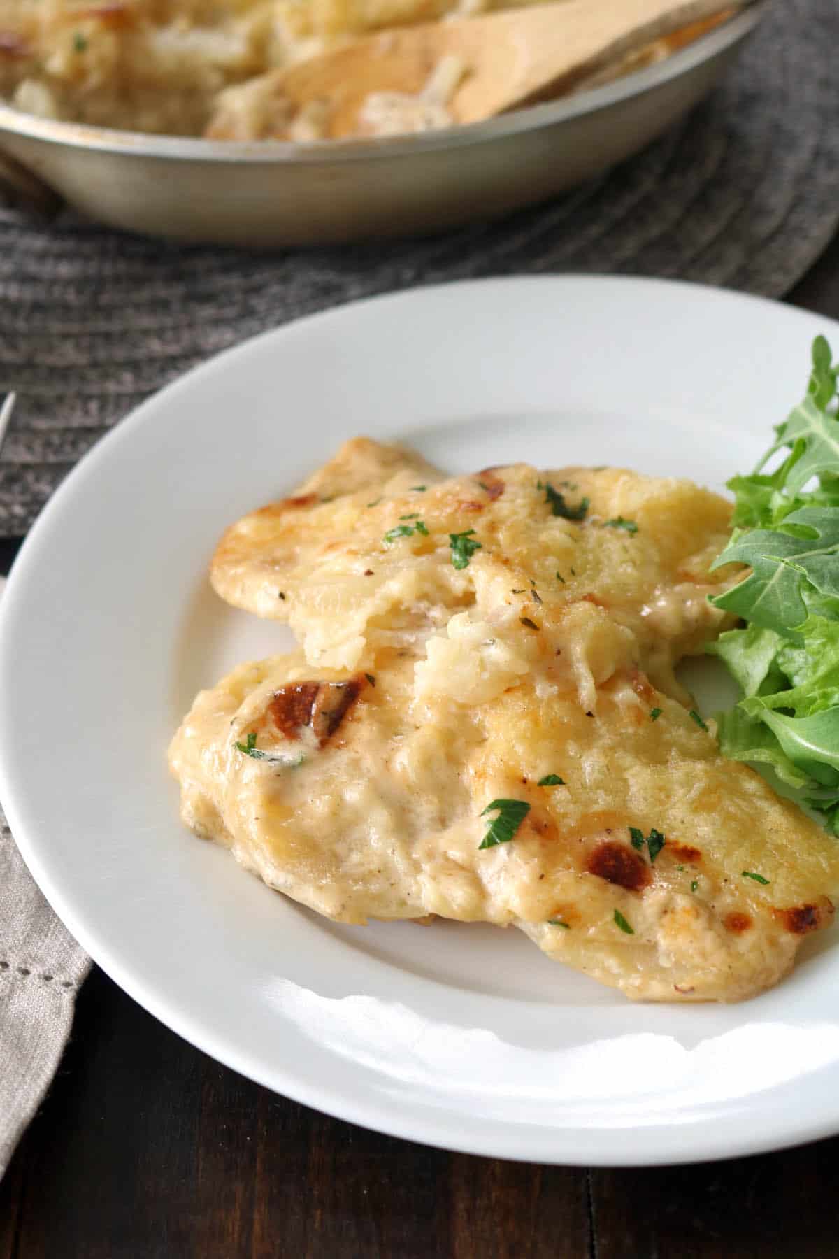 A white plate with two pieces of creamy baked fish, garnished with herbs, served alongside a small portion of fresh arugula greens. A skillet with more fish is visible in the background.