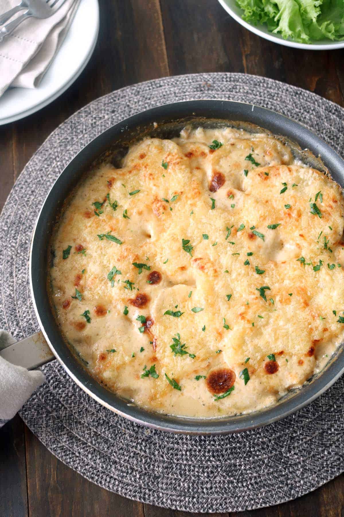 A skillet filled with creamy, baked gratin topped with melted, golden-brown cheese and sprinkled with chopped herbs, sits on a gray placemat on a wooden table.