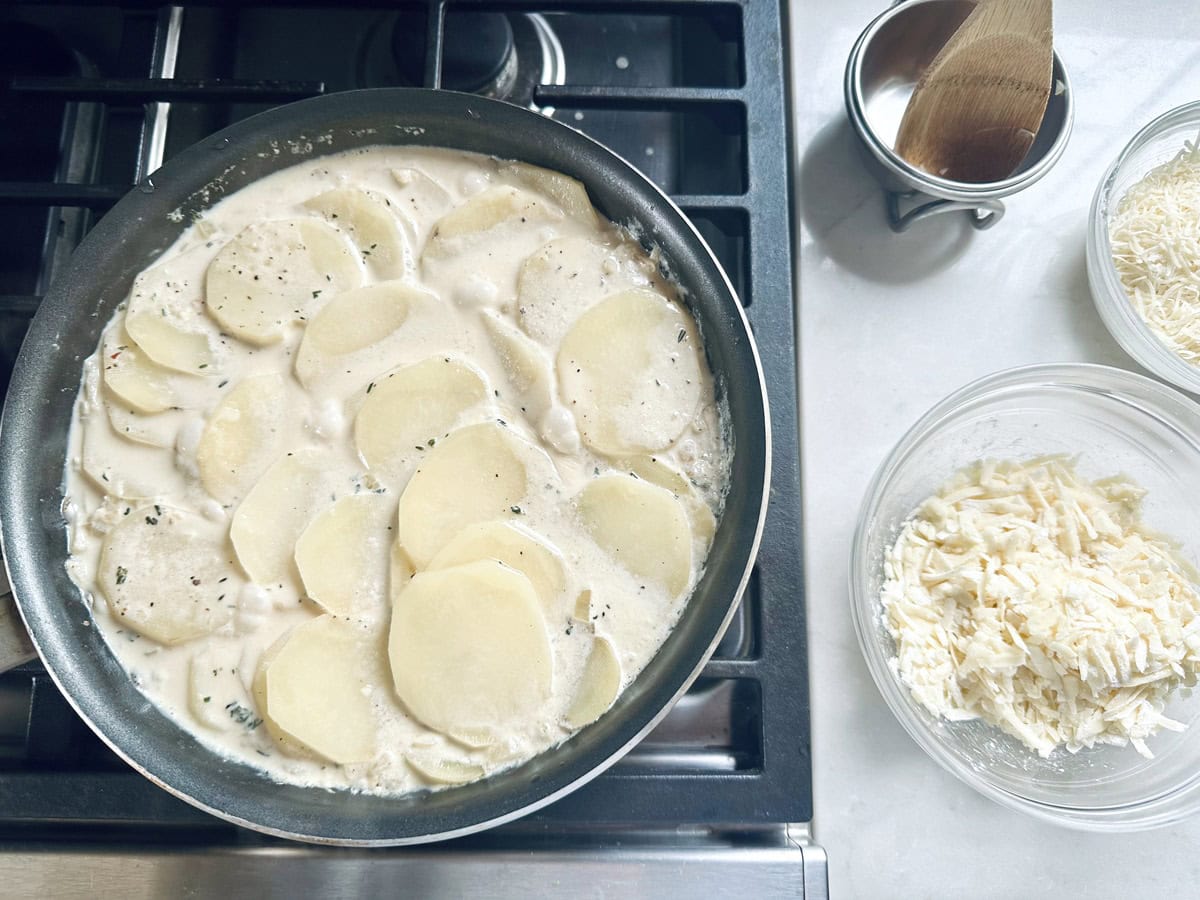 A pan on a stove contains sliced potatoes in a creamy sauce. Next to it are bowls with shredded cheese and a wooden spoon in a metal cup on a white countertop.
