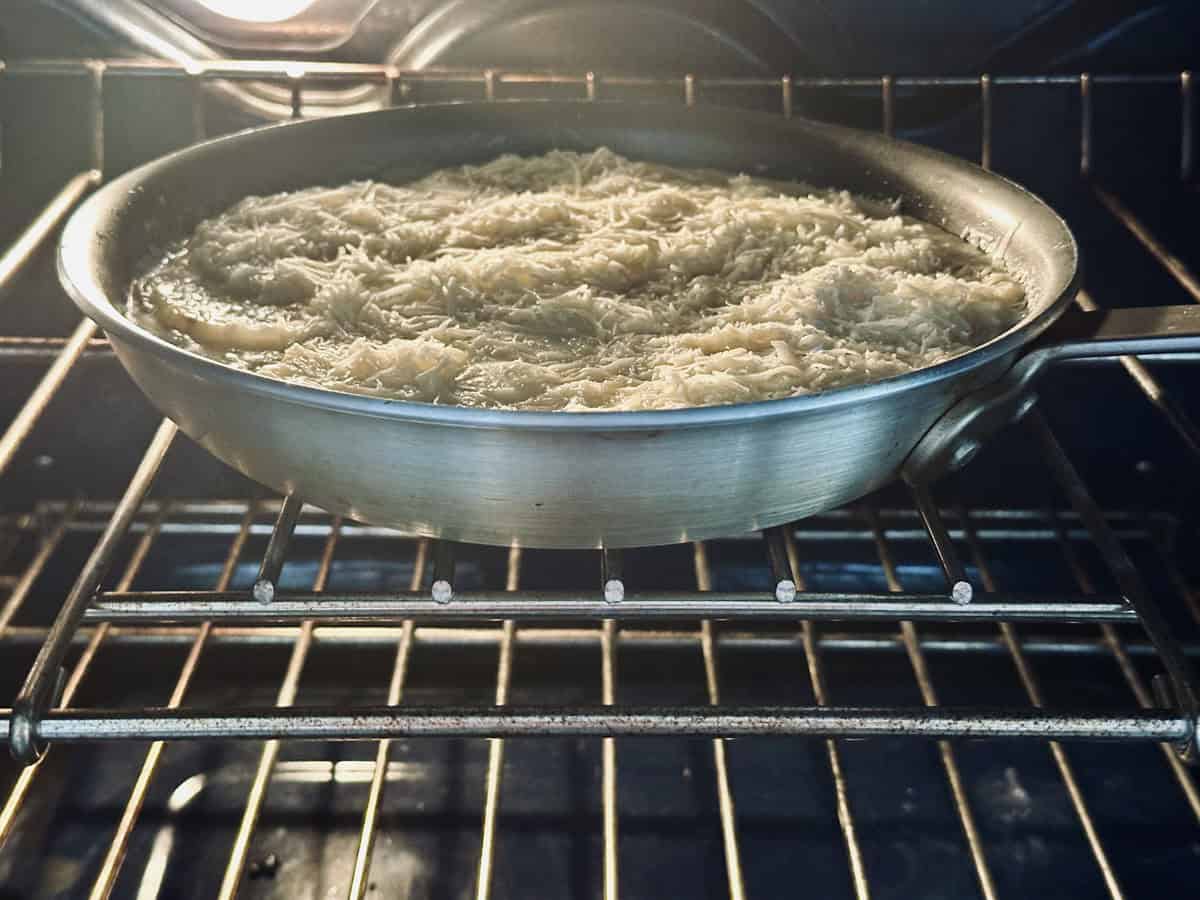 A round metal pan filled with shredded potatoes is baking on the middle rack of an oven, illuminated by the oven light.