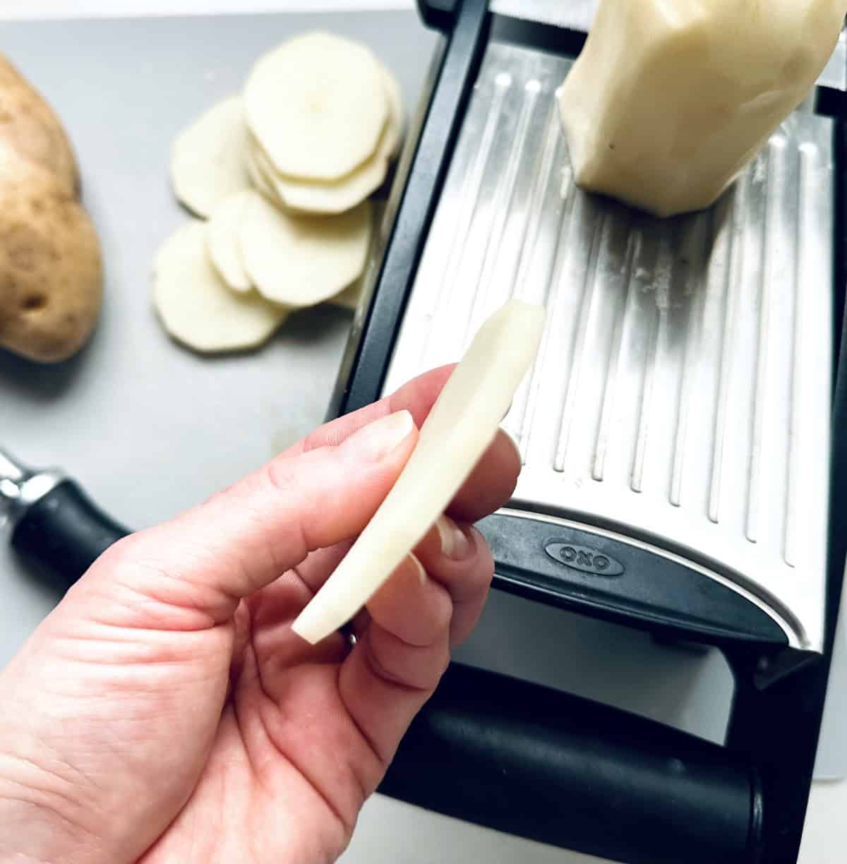 A hand holds a thinly sliced potato piece above an OXO mandoline slicer, with more potato slices and a whole potato visible on a cutting board in the background.