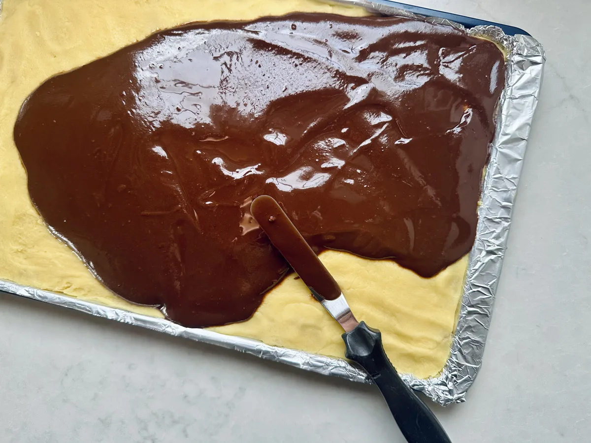 A baking tray lined with foil holds a yellow dough base being spread with glossy chocolate icing using an offset spatula.
