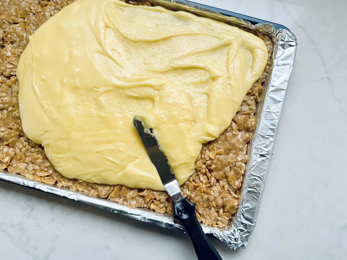 A baking tray lined with foil holds a layer of oat and peanut butter mixture being topped with creamy yellow frosting, spread with an offset spatula on a white countertop.