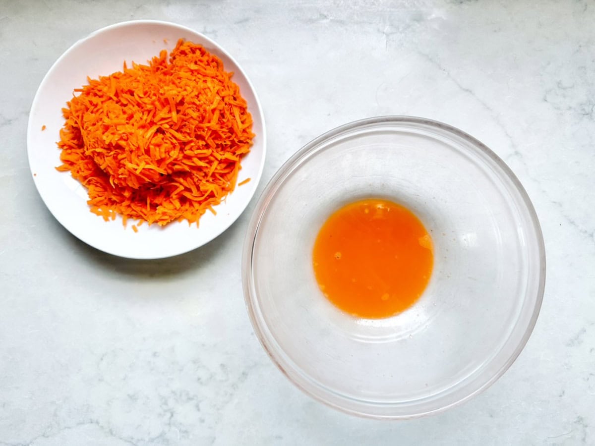 A white bowl filled with shredded carrots sits next to a clear glass bowl containing a small amount of orange carrot juice on a light-colored surface.