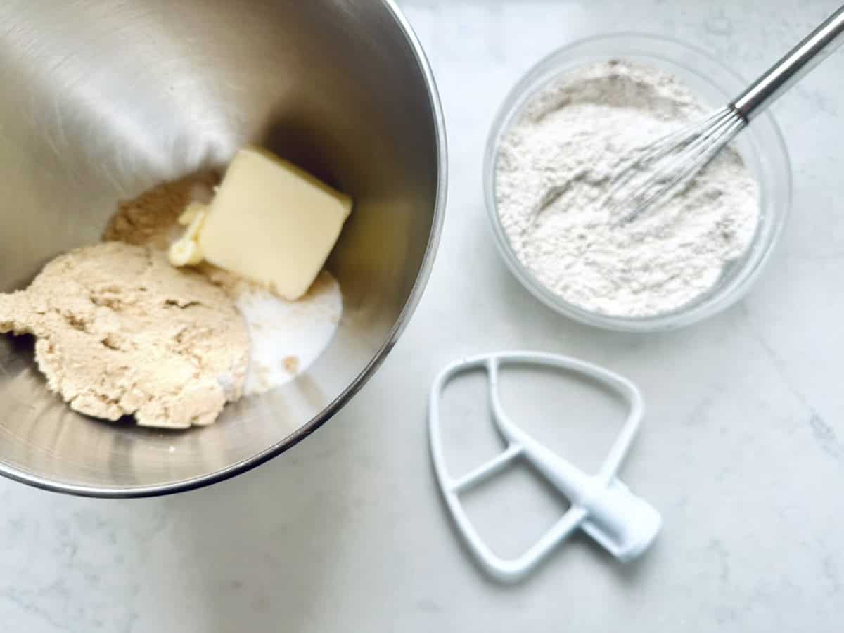 A mixing bowl with brown sugar, white sugar, and a chunk of butter sits next to a bowl of flour with a whisk, and a paddle mixer attachment on a white surface.