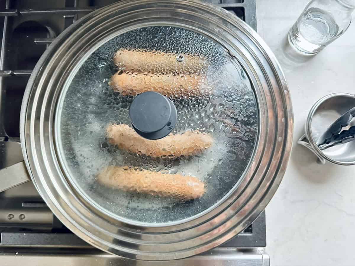Three sausages cook in a pan under a glass lid, steam gathering as you prep arugula salad ingredients for quick salad meals nearby. A glass of water, metal tongs, and a small container rest on the white countertop.