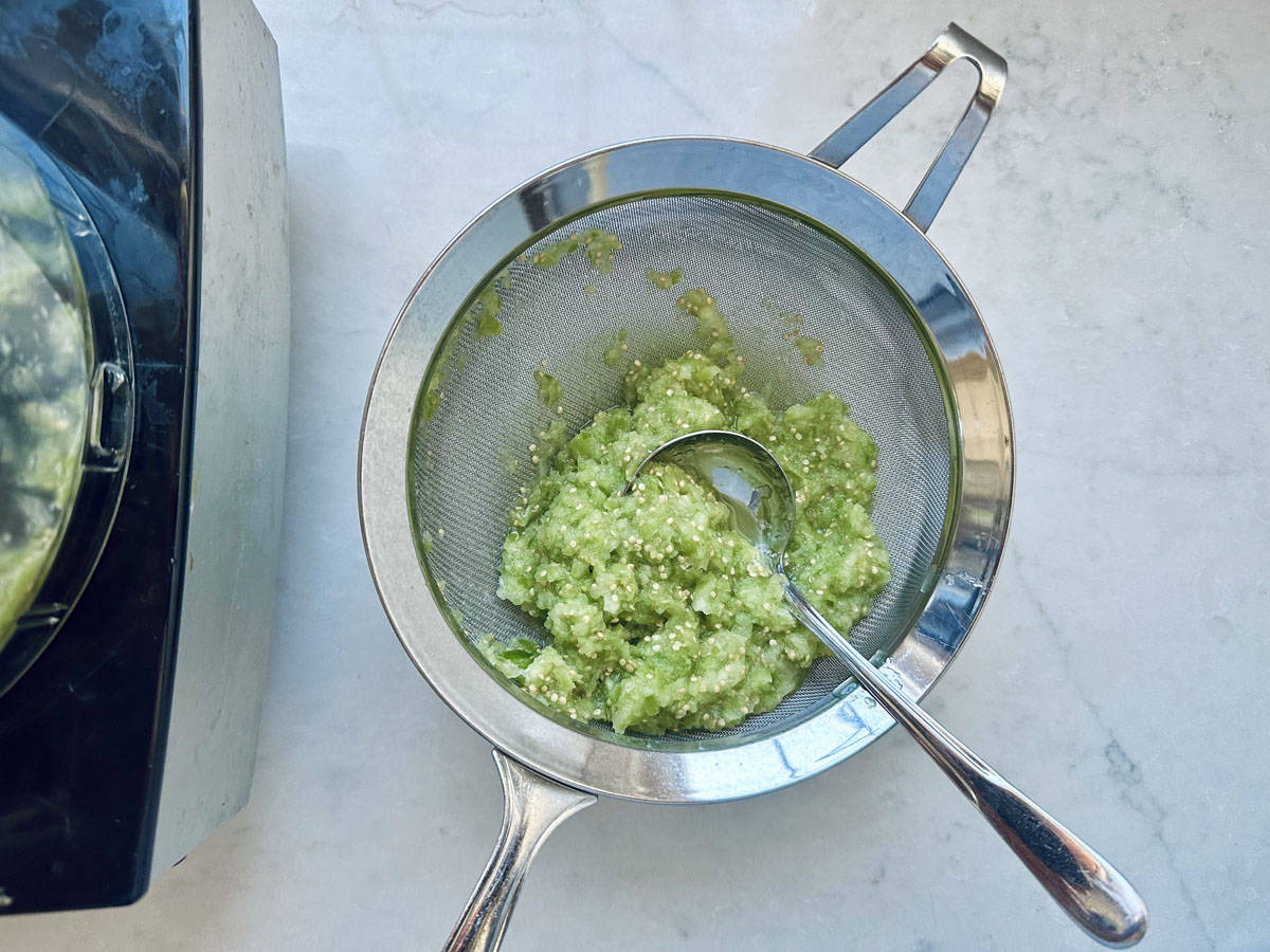 A metal strainer containing blended green mixture sits over a bowl on a white countertop, with a spoon resting inside. A food processor is partially visible on the left.