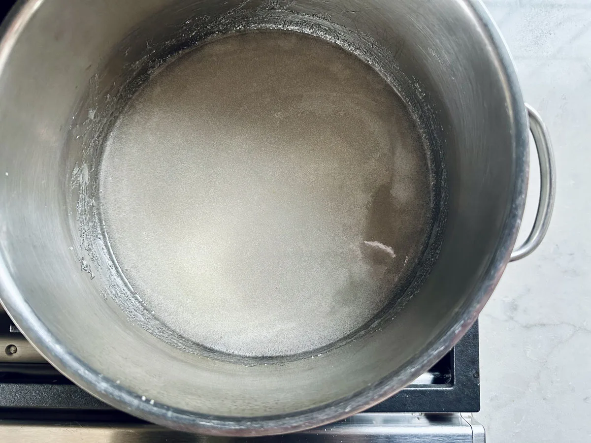 A large metal pot filled with a thin layer of clear water and undissolved sugar sits on a stovetop, viewed from above. The sugar is visible as a white, grainy layer at the bottom of the pot.