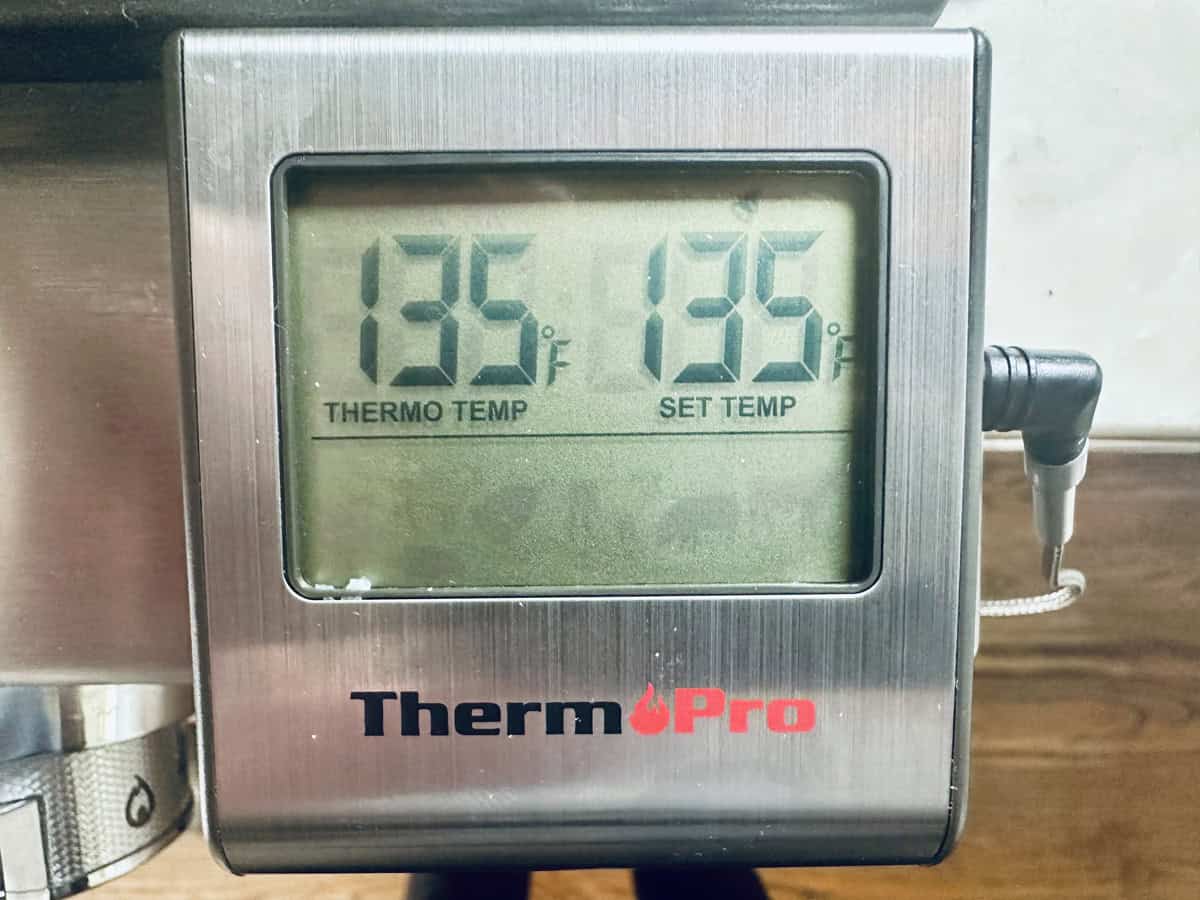 A digital ThermoPro thermometer displays both the current and set temperatures at 135°F while monitoring a Crumb-Crusted Rack of Lamb Persillade. A metal probe is plugged into the side, and the device rests on a metal surface.