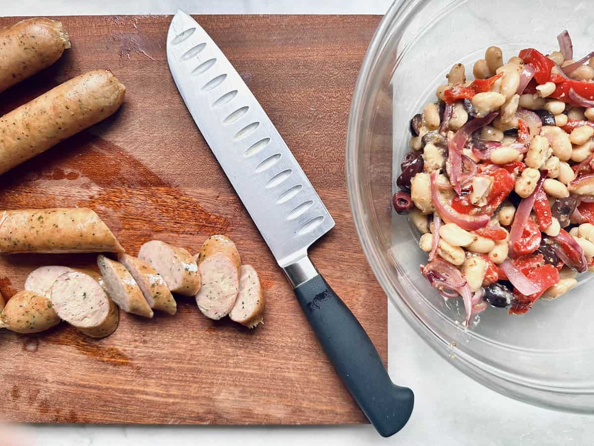 A sliced sausage on a wooden cutting board next to an unsliced sausage and a chef’s knife, with a glass bowl of quick salad meal featuring classic arugula salad ingredients like beans, olives, and red peppers nearby.