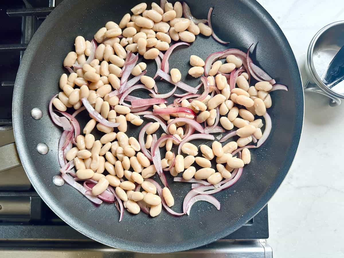 A black skillet on a stove contains white beans and thinly sliced red onions being sautéed—a perfect base for quick salad meals like Arugula Salad with Sausage and White Beans. A metal measuring cup rests on the counter to the right.