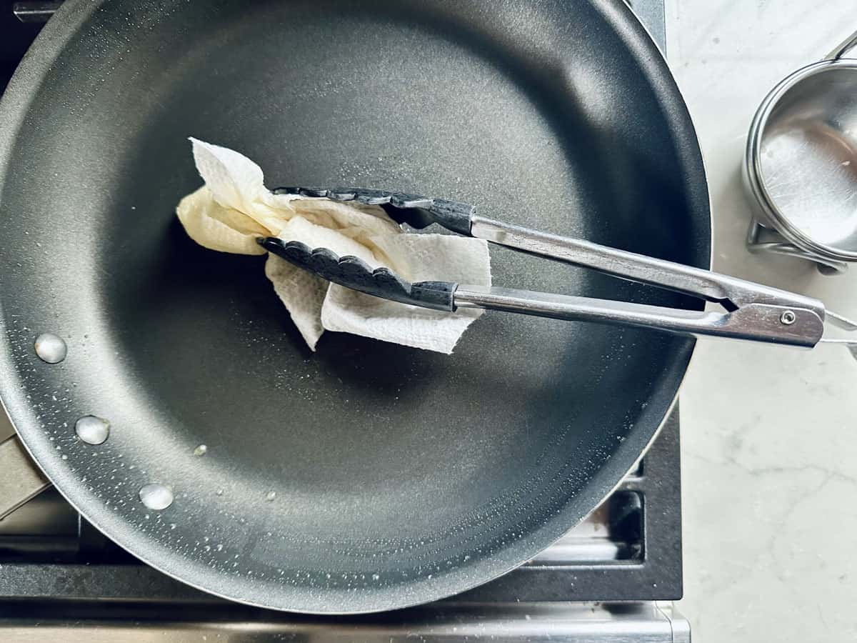 A pair of metal tongs holds a folded paper towel, wiping the surface of a nonstick frying pan—perfect prep for making quick salad meals like Arugula Salad with Sausage and White Beans. A small saucepan sits nearby on a light-colored countertop.