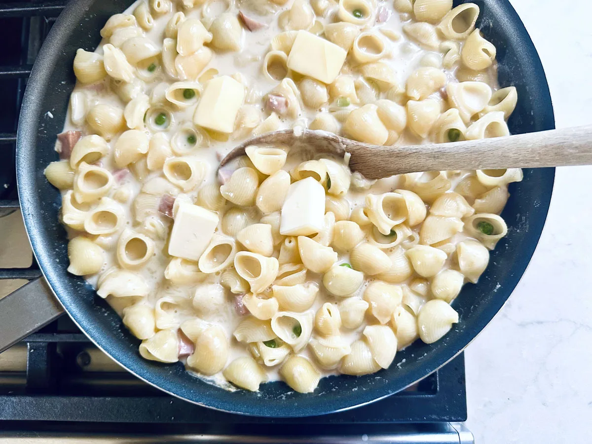 A skillet filled with creamy shell pasta, diced ham, green onions, and several cubes of butter, being stirred with a wooden spoon on a stovetop.