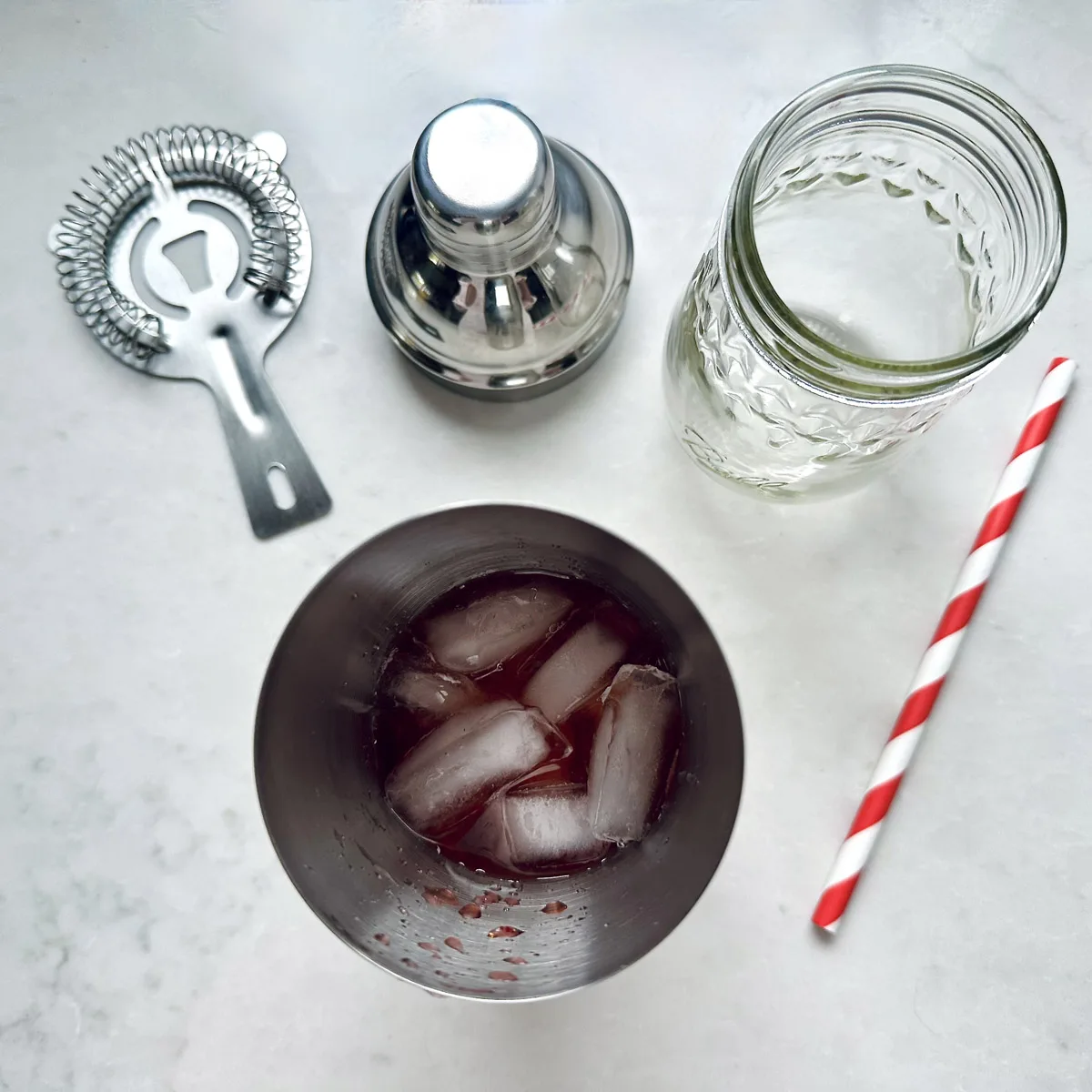 A cocktail shaker with ice and red liquid, a metal strainer, an empty patterned glass jar, and a red-and-white striped paper straw on a white surface.