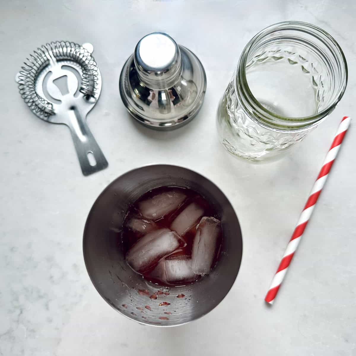A cocktail shaker with ice and red liquid, a metal strainer, an empty patterned glass jar, and a red-and-white striped paper straw on a white surface.