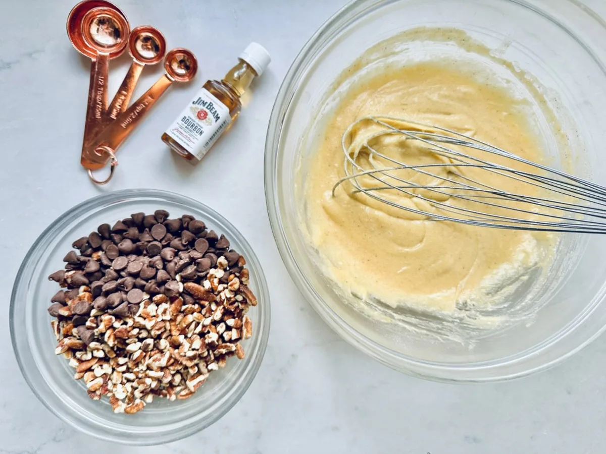 A bowl of batter being whisked sits next to a bowl of chocolate chips and chopped nuts, a set of copper measuring spoons, and a small bottle of vanilla extract on a white surface.