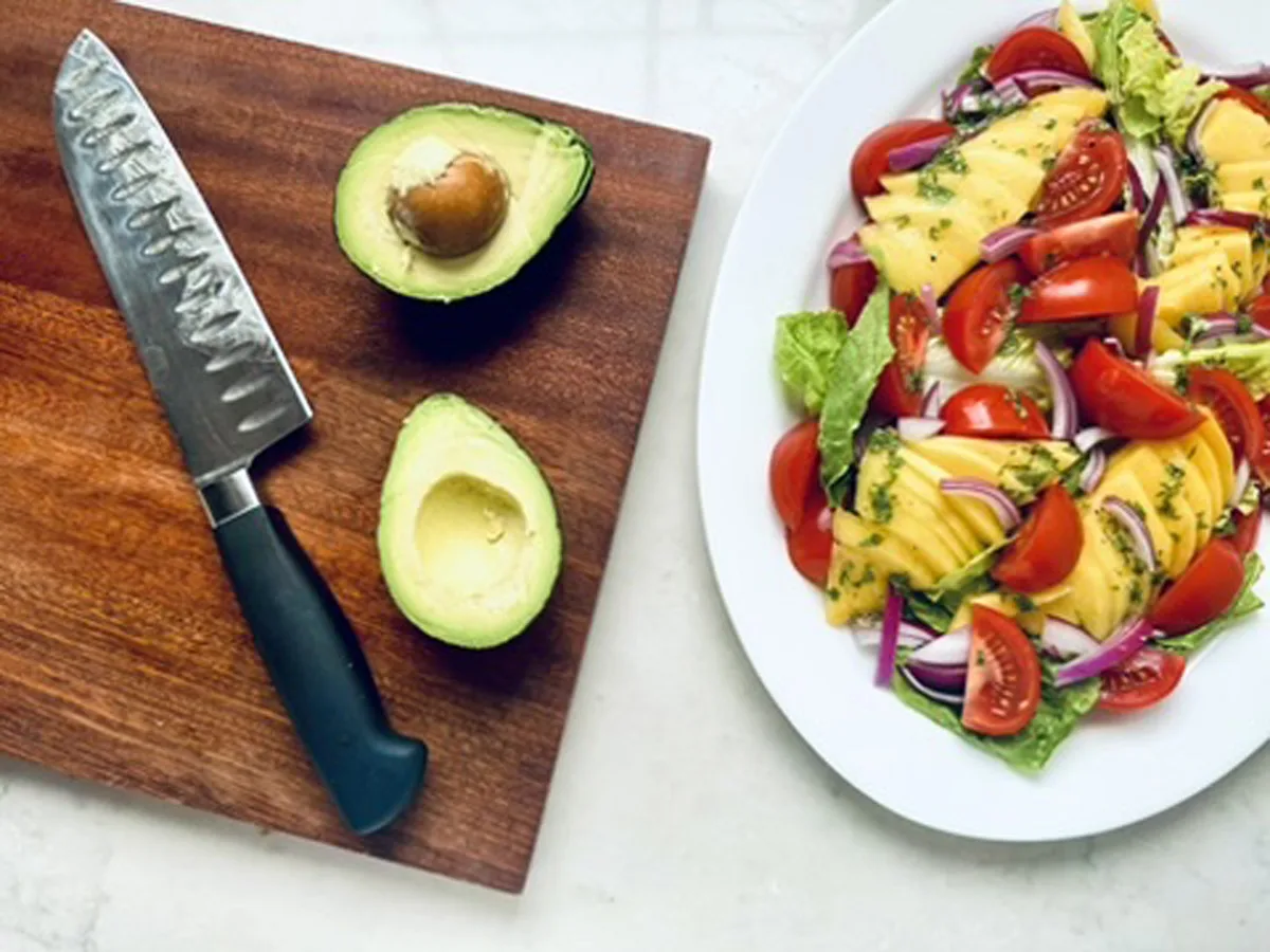 A halved avocado and a knife rest on a wooden cutting board next to a plate of Mango and Avocado Salad with cherry tomatoes, red onion, and greens on a white surface.