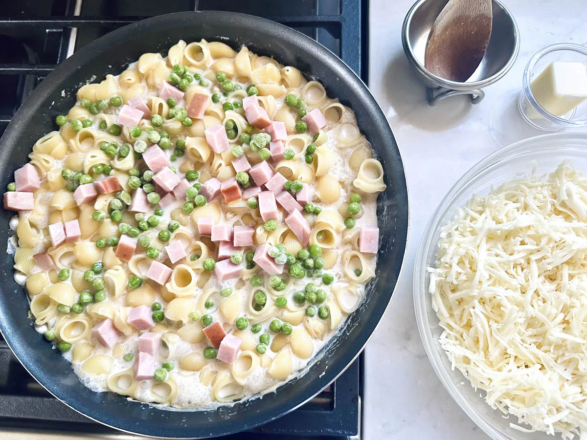 A skillet on a stove contains pasta shells, peas, and cubed ham in a creamy sauce. Next to the skillet are a bowl of shredded cheese, a small dish of butter, and a wooden spoon resting in a metal cup.