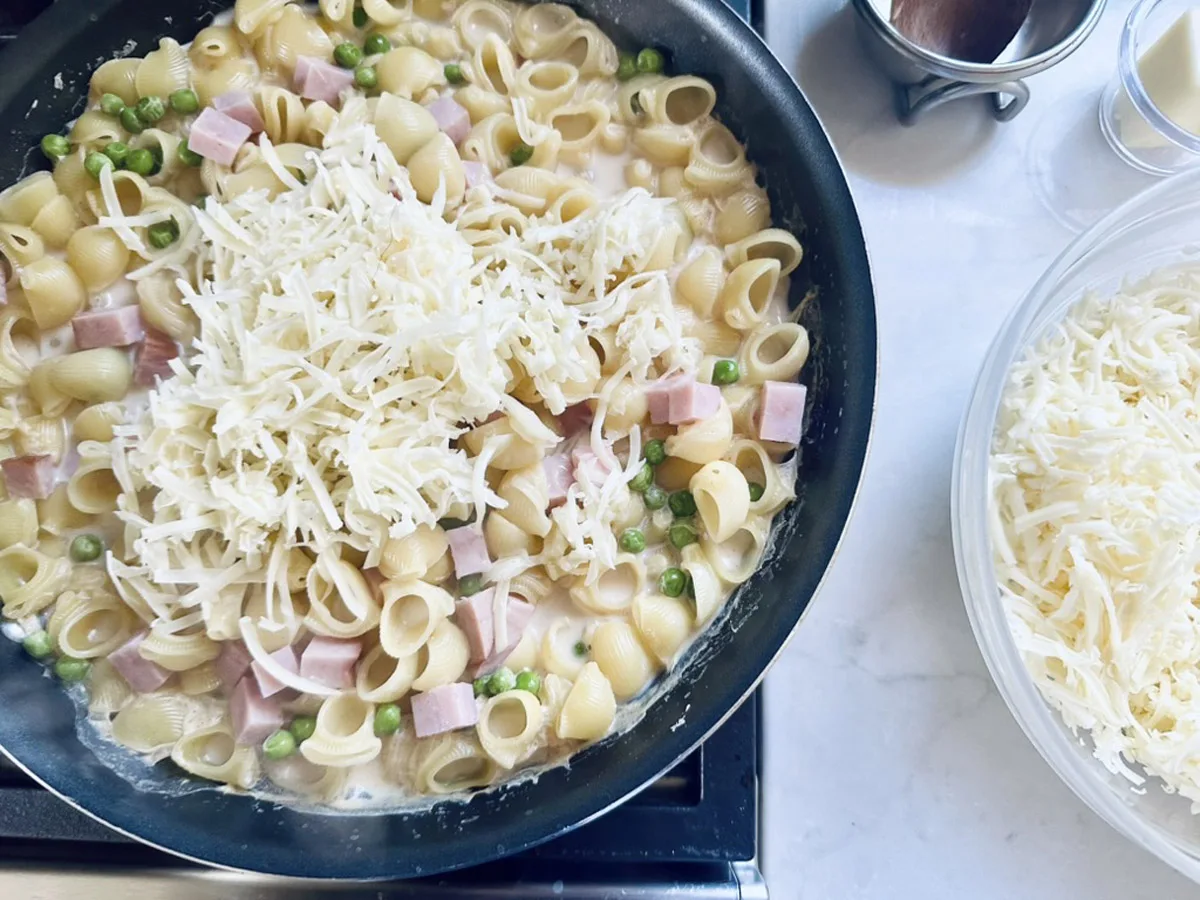 A skillet filled with shell pasta, green peas, and ham cubes in a creamy sauce, topped with shredded cheese, sits on a stovetop next to a bowl of more cheese on a white countertop.